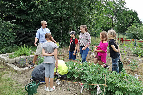 Im Schulgarten: Sechs Grundschüler hören einem jungen Mann zu, der etwas über Gartenarbeit erklärt.