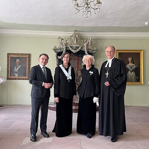 Mann in Anzug, zwei Frauen in Äbtissinnentracht, Pastor im Talar in Saal mit Gemälden und Kronleuchter
