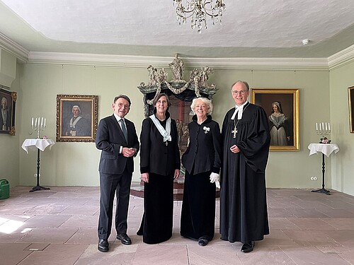 Mann in Anzug, zwei Frauen in Äbtissinnentracht, Pastor im Talar in Saal mit Gemälden und Kronleuchter