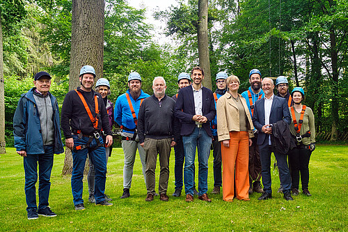 Gruppenbild mit 13 Personen vor hohen Bäumen. Einige tragen Helm und Klettersteiggurt.
