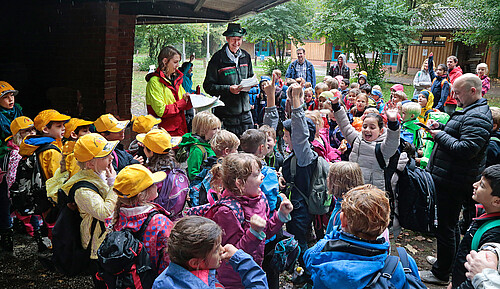 Eine große Gruppe von Grundschülern steht im Regen neben einer Holzhütte und freut sich über Urkunden eines Wettbewerbs im Wald.
