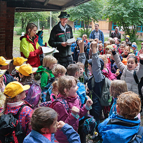 Eine große Gruppe von Grundschülern steht im Regen neben einer Holzhütte und freut sich über Urkunden eines Wettbewerbs im Wald.
