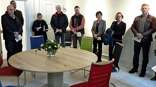 Acht Personen stehen in einem hell gestalteten Raum hinter einem hölzernen Tisch.