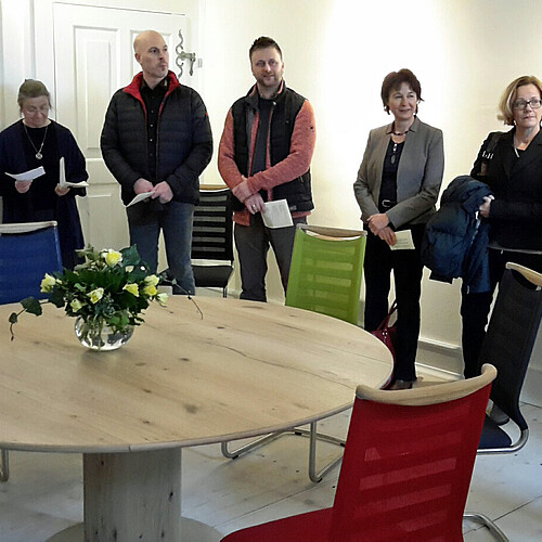 Acht Personen stehen in einem hell gestalteten Raum hinter einem hölzernen Tisch.