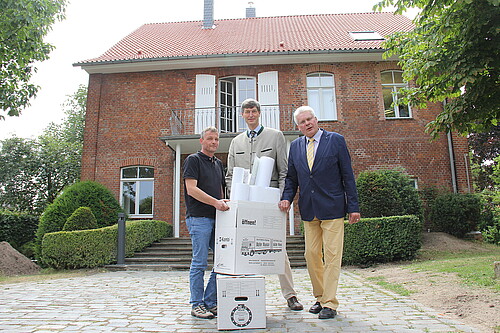 Drei Herren haben zwei Umzugskartons vor sich. Sie stehen vor einem Haus mit Backsteinfassade.