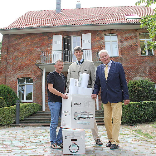 Drei Herren haben zwei Umzugskartons vor sich. Sie stehen vor einem Haus mit Backsteinfassade.