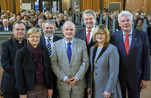 Fünf Männer und zwei Frauen stehen in einer Gruppe vor gefüllten Bänken in einer Kirche.
