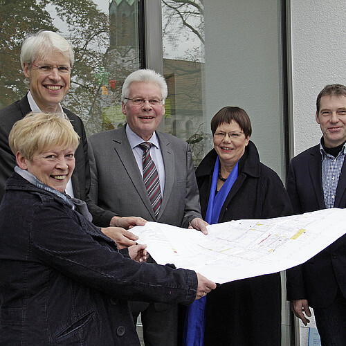 Zwei Frauen und drei Männer stehen in einem Halbkreis vor einer modernen Glasfassade und halten einen Bauplan in den Händen.