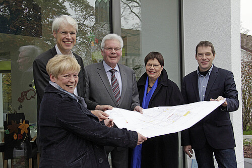 Zwei Frauen und drei Männer stehen in einem Halbkreis vor einer modernen Glasfassade und halten einen Bauplan in den Händen.