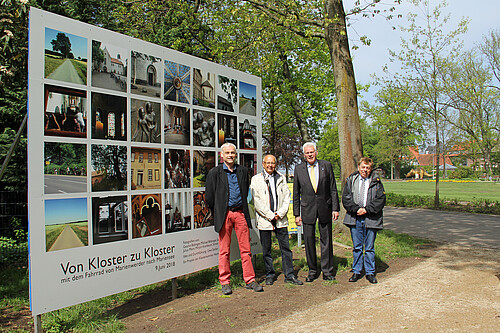 Drei Personen stehen vor einer drei mal vier Meter großen Tafel, auf der 28 quadratische Fotos zu sehen sind.