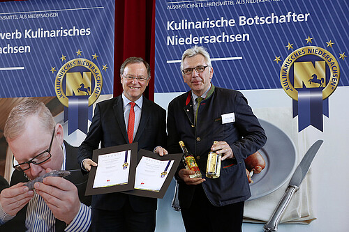 Zwei Männer stehen auf einer Bühne. Einer der hält zwei Urkunden in der Hand, der andere zwei Flaschen mit Spirituosen.