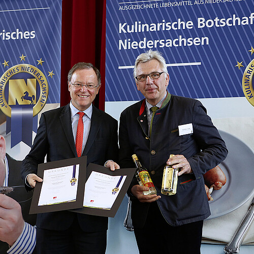 Zwei Männer stehen auf einer Bühne. Einer der hält zwei Urkunden in der Hand, der andere zwei Flaschen mit Spirituosen.