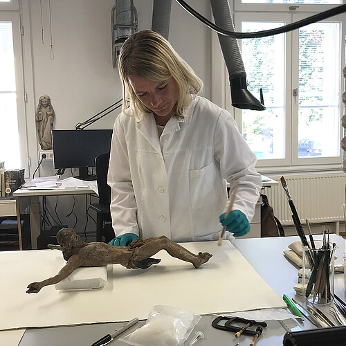 Eien Frau im weißen Laborkittel steht hinter einem Arbeitstisch, auf dem eine hölzerne Jesus-Figur liegt.