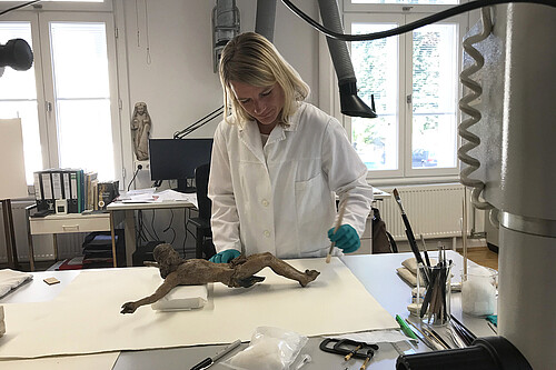 Eien Frau im weißen Laborkittel steht hinter einem Arbeitstisch, auf dem eine hölzerne Jesus-Figur liegt.