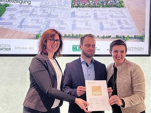 Zwei Frauen und ein Mann halten eine Urkunde vor einer Leinwand