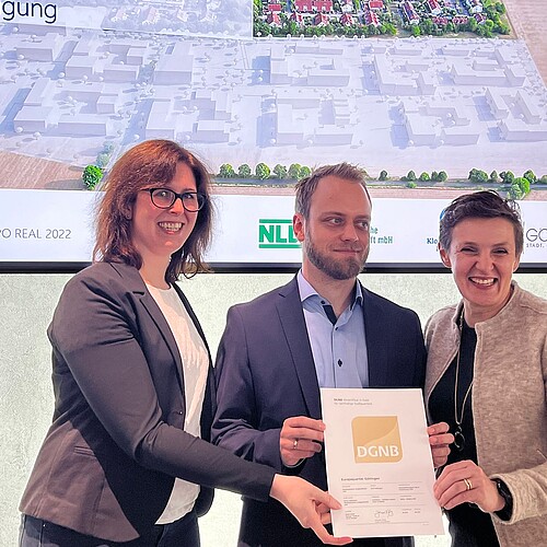 Zwei Frauen und ein Mann halten eine Urkunde vor einer Leinwand