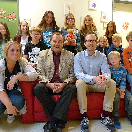 Kinder aus dem Geschwisterclub sitzen mit Betreuerin und Förderern auf einem Sofa und schauen in die Kamera.