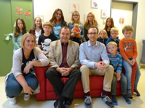 Kinder aus dem Geschwisterclub sitzen mit Betreuerin und Förderern auf einem Sofa und schauen in die Kamera.