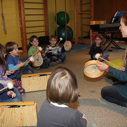 Sieben Kleinkinder und zwei Erzieherinnen, der Evangelisch-lutherischen Kindertagesstätte Nazareth, sitzen auf dem Fußboden und trommeln.