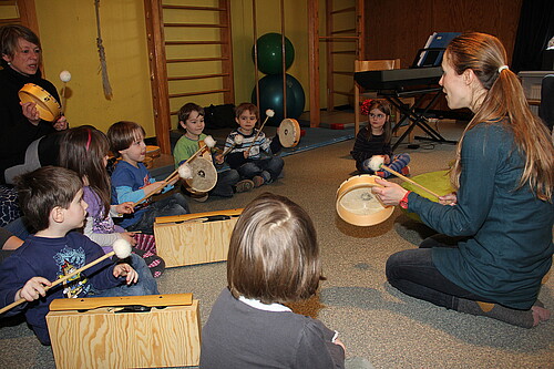 Sieben Kleinkinder und zwei Erzieherinnen, der Evangelisch-lutherischen Kindertagesstätte Nazareth, sitzen auf dem Fußboden und trommeln.