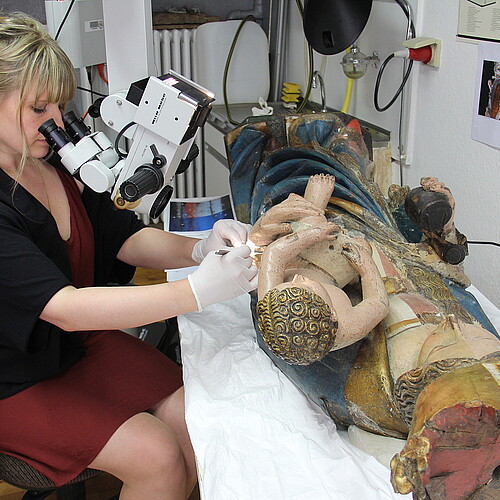 Eine Frau schaut durch ein Spezialgerät für Restauratoren auf eine vor ihr liegende Holzfigur, an der sie gerade arbeitet.