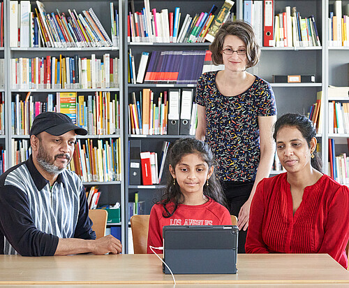 Zwei Erwachsene und ein Schulkind sitzen an einem Tisch vor einem Tablet, dahinter steht eine Frau.