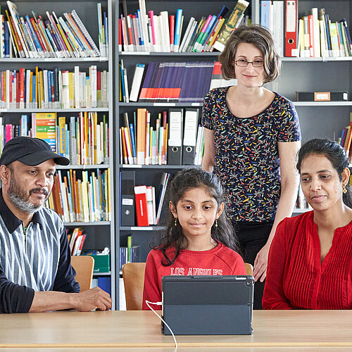 Zwei Erwachsene und ein Schulkind sitzen an einem Tisch vor einem Tablet, dahinter steht eine Frau.