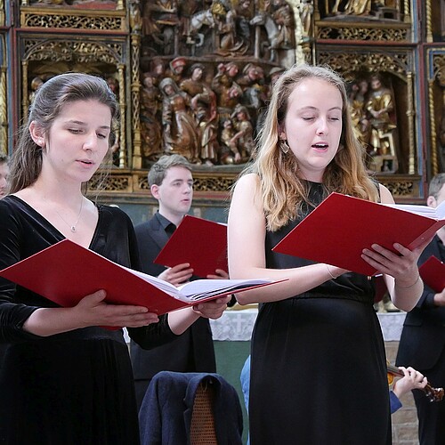 Zwei junge Frauen und drei junge Männer stehen vor einem geschnitzten Altaraufsatz und singen.
