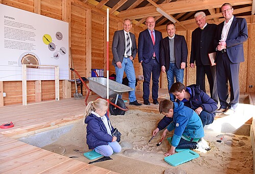 Fünf Herren stehen hinter einem mit Sand gefüllten Quader, in welchen drei Kinder graben.