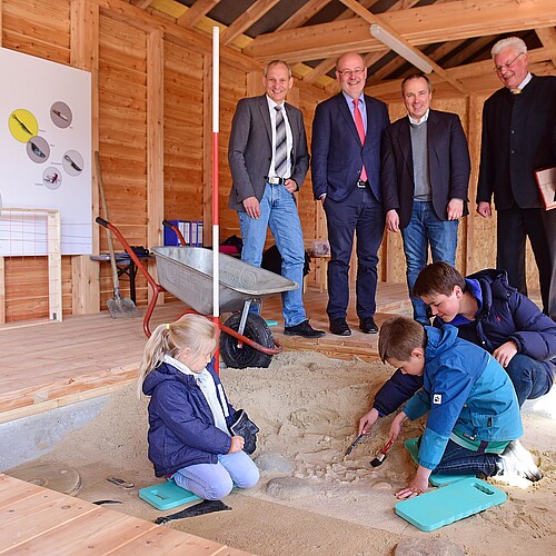 Fünf Herren stehen hinter einem mit Sand gefüllten Quader, in welchen drei Kinder graben.