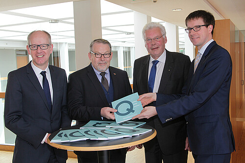 Vier Herren stehen hinter einen Stehtisch nebeneinander. Sie halten eine Broschüre in den Händen.