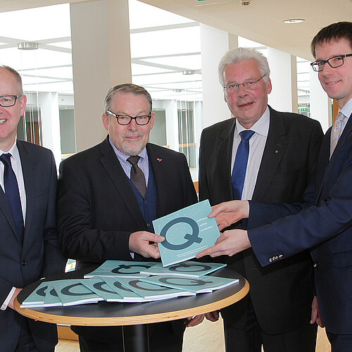 Vier Herren stehen hinter einen Stehtisch nebeneinander. Sie halten eine Broschüre in den Händen.