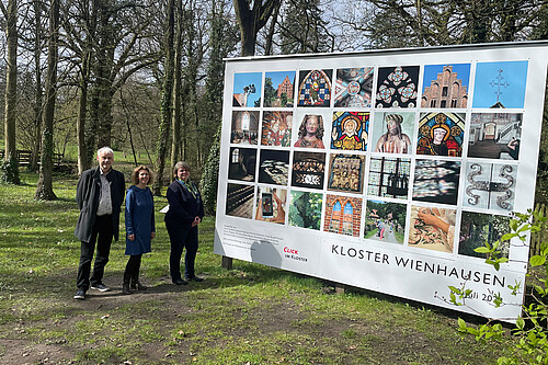 Drei Personen stehen neben einer großformatigen Tafel mit 28 quadratischen Fotos.