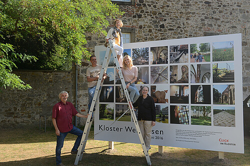 Drei Frauen und zwei Männer stehen vor einer Bild-Tafel.