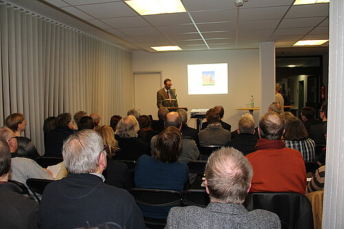 Eine Gruppe von Personen hört einem Redner am Pult zu.