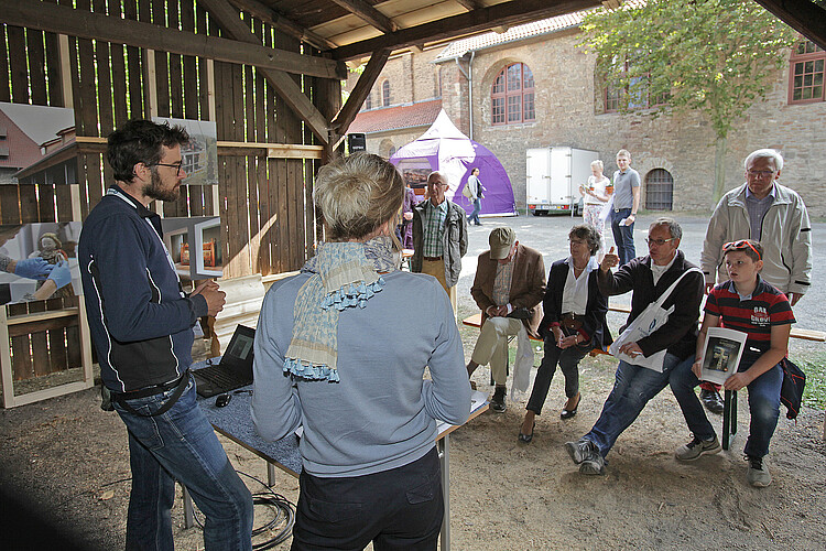 Klosterkammerfest 2018 Zwei Personen stehen in einem Unterstand und halten einen Vortrag, davor sitzen mehrer Personen auf einer Bank und hören zu.