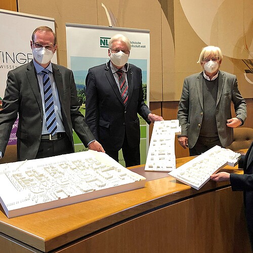 Vier Männer mit Mund-Nase-Masken stehen um einen Tisch herum und präsentieren Architektur-Modelle.