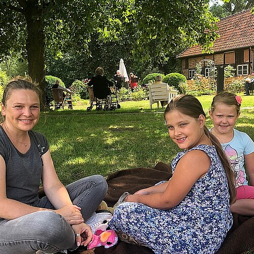 Eine Frau und zwei Mädchen auf einer Decke im Garten.