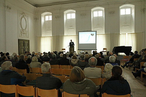 In Stuhlreihen sitzen Menschen, auf einem Podium steht eine Person die zu ihnen spricht.
