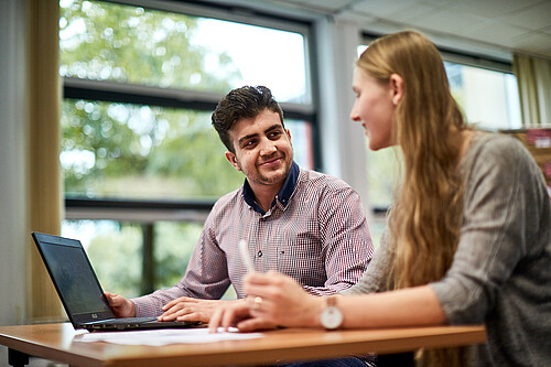 Zwei Jugendliche sitzen zusammen an einem Tisch und sehen sich an, vor ihnen steht ein Laptop.