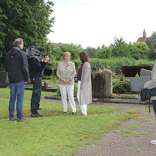 Zwei Frauen stehen auf einer Rasenfläche und unterhalten sich. Sie werden dabei von einem dreiköpfigen Kamerateam gefilmt.