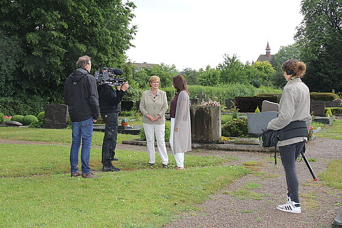 Zwei Frauen stehen auf einer Rasenfläche und unterhalten sich. Sie werden dabei von einem dreiköpfigen Kamerateam gefilmt.