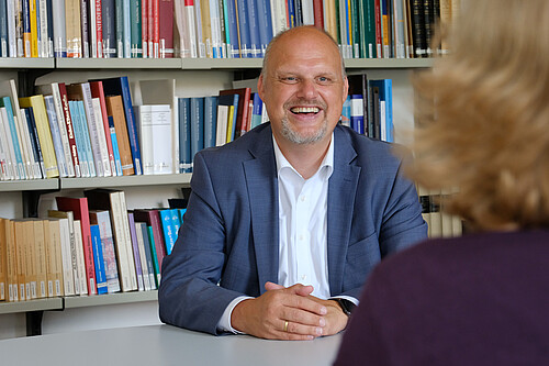 Ein Mann in blauen Jackett und weißen Hemd sitzt vor Bücherregalen.