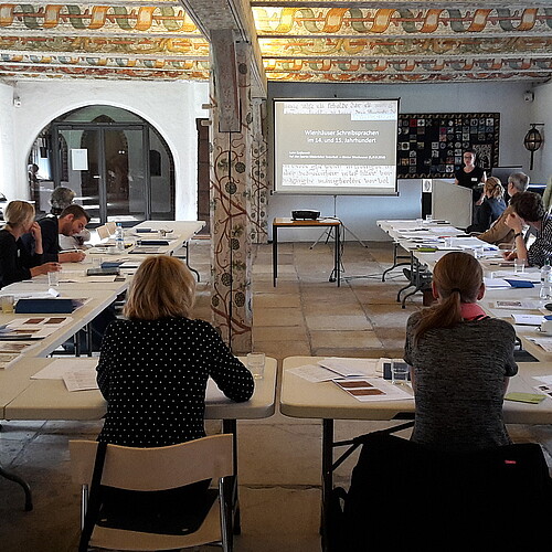 In einem mit Raum mit kunstvoll bemahlten Deckenbalken im Kloster Wienhausen sitzt eine Gruppe von rund 15 Personen zusammen.
