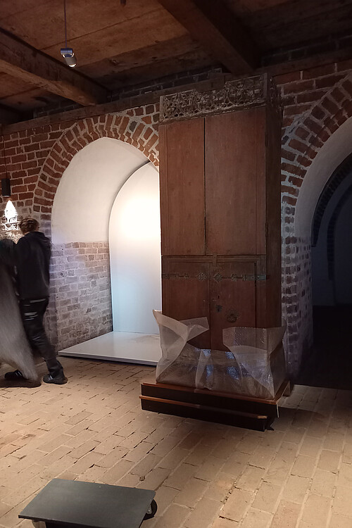 Ein hoher, hölzerner Schrank zwischen zwei Torbögen wurde auf ein Podest aus Holz gestellt. Im unteren Bereich ist er mit Plastikfolie eingeschlagen.