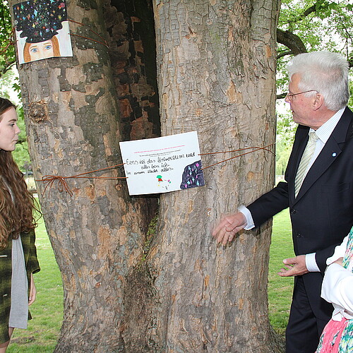 Drei Erwachsene und eine Schülerin stehen an beiden Seiten von einem Baumstamm, den die Schülerin mit einem Kunstwerk verziert hat.