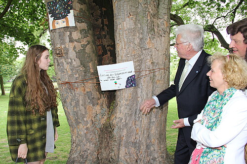 Drei Erwachsene und eine Schülerin stehen an beiden Seiten von einem Baumstamm, den die Schülerin mit einem Kunstwerk verziert hat.