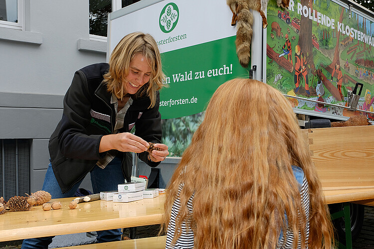Stefanie Grevelhörster, Revierleiterin und Waldpädagogin der Klosterforsten, im Einsatz neben dem Waldmobil zu sehen. Sie steht hinter einen Tisch, davor steht ein Mädchen, dass von hinten zu sehen ist.