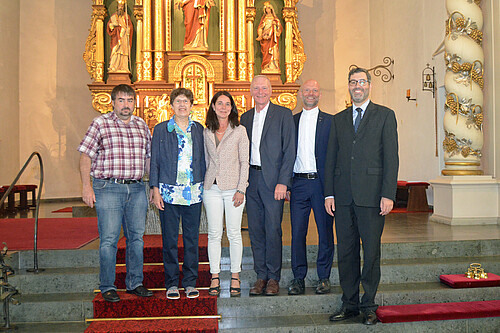 Zwei Frauen und vier Männer stehen im Innenraum einer Kirche auf einer Treppe.
