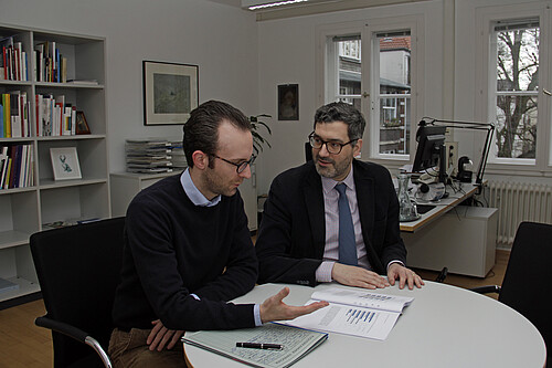 Zwei Männer sitzen an einem Tisch in einem Arbeitsraum, auf dem Tisch liegen Unterlagen.